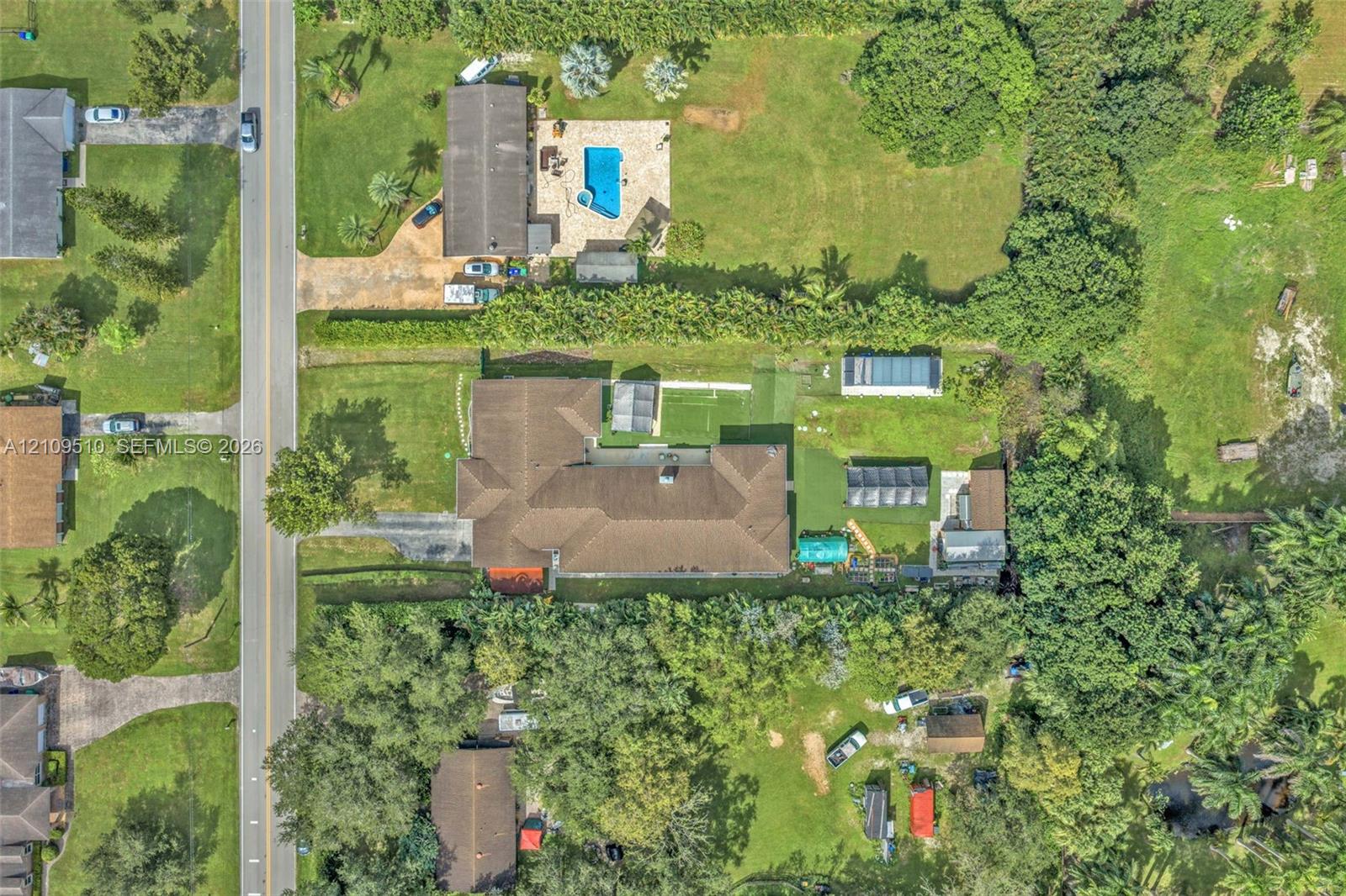 Aerial perspective of a sprawling property featuring extensive grass lawn, mature perimeter trees, a multi-section roofline, and paved driveway