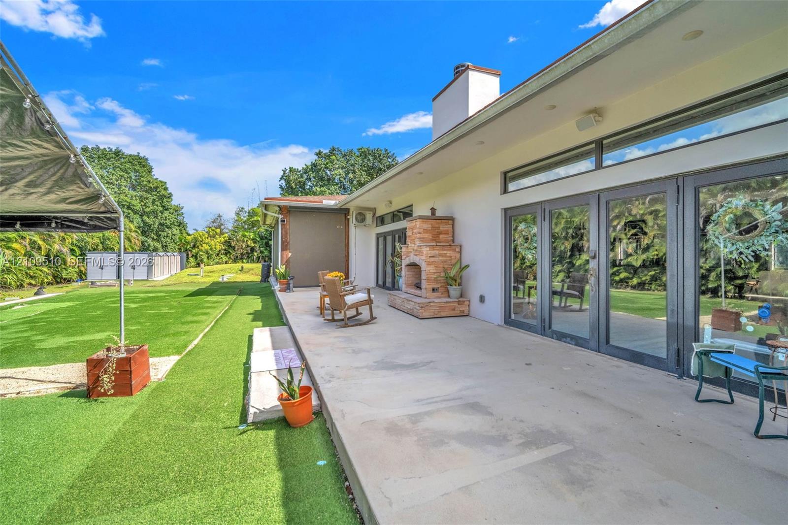 Spacious concrete patio featuring a built-in masonry outdoor fireplace, large glass multi-panel doors, and direct access to an expansive synthetic turf lawn