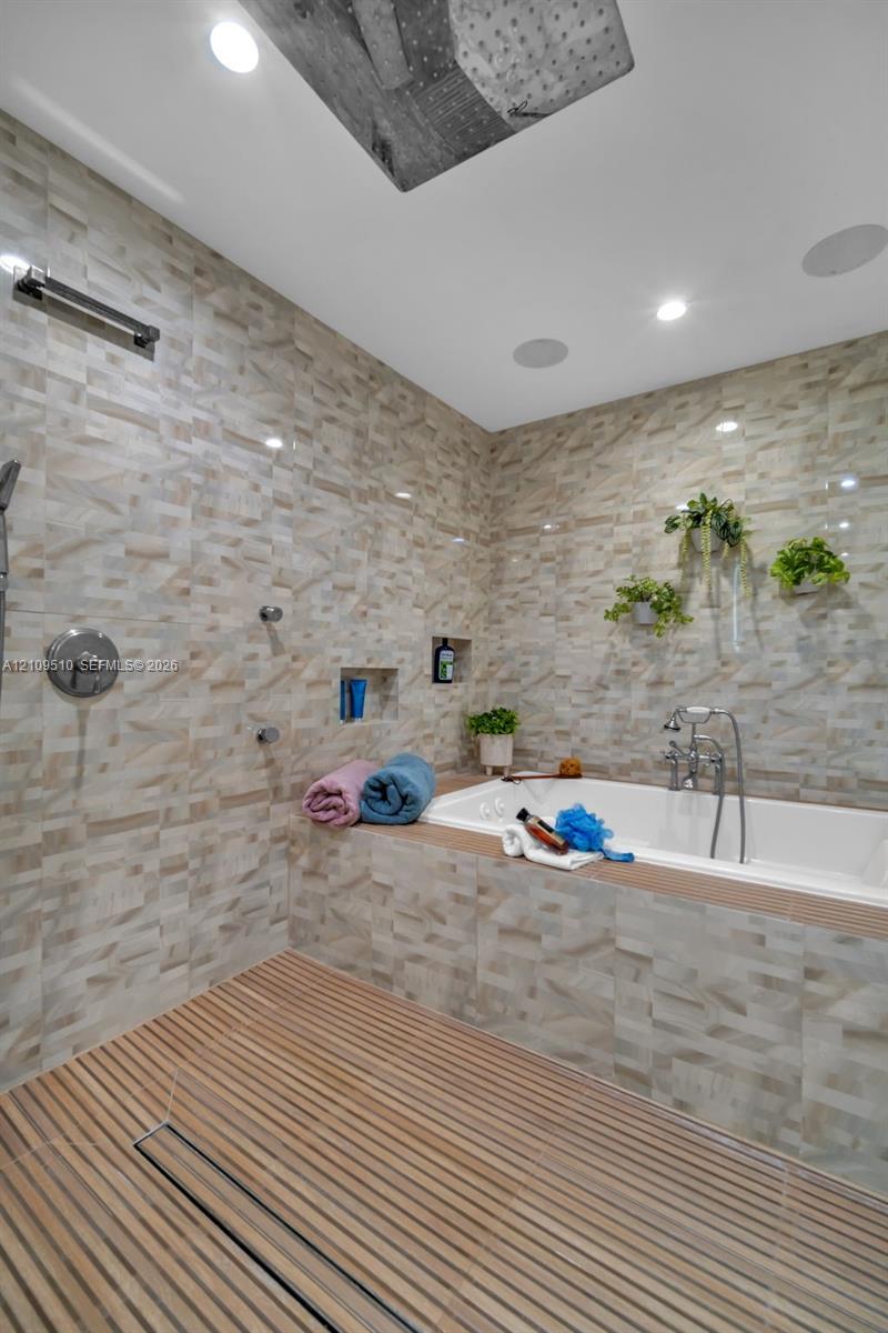 Wet room featuring a deep soaking tub, ceiling-mounted rainfall showerhead, linear floor drain, wood-slat flooring, and textured tile wall cladding