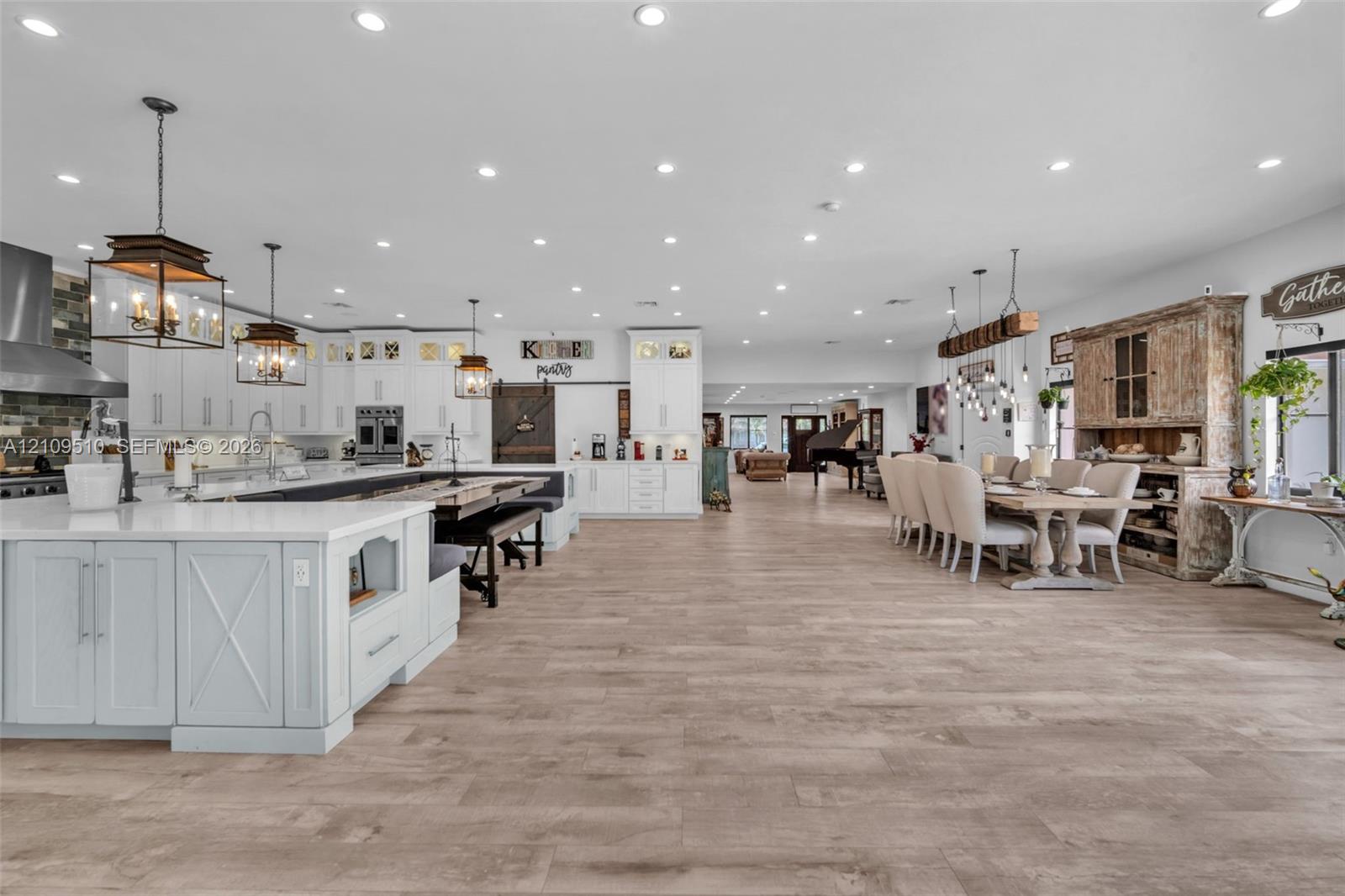 Open-concept space featuring wood-finish flooring, extensive recessed lighting, white shaker-style cabinetry, a large kitchen island with quartz countertops, and three oversized metal-frame pendant lights