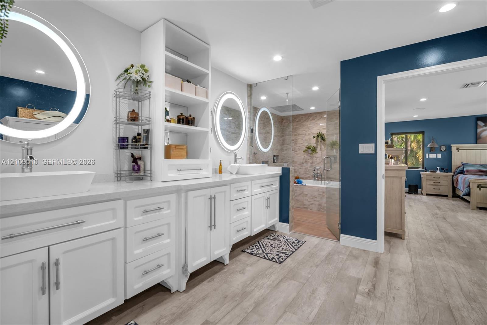 Dual white vanities featuring vessel sinks, integrated shelving, lighted circular mirrors, and a walk-in glass-enclosed shower with stone-tile surround