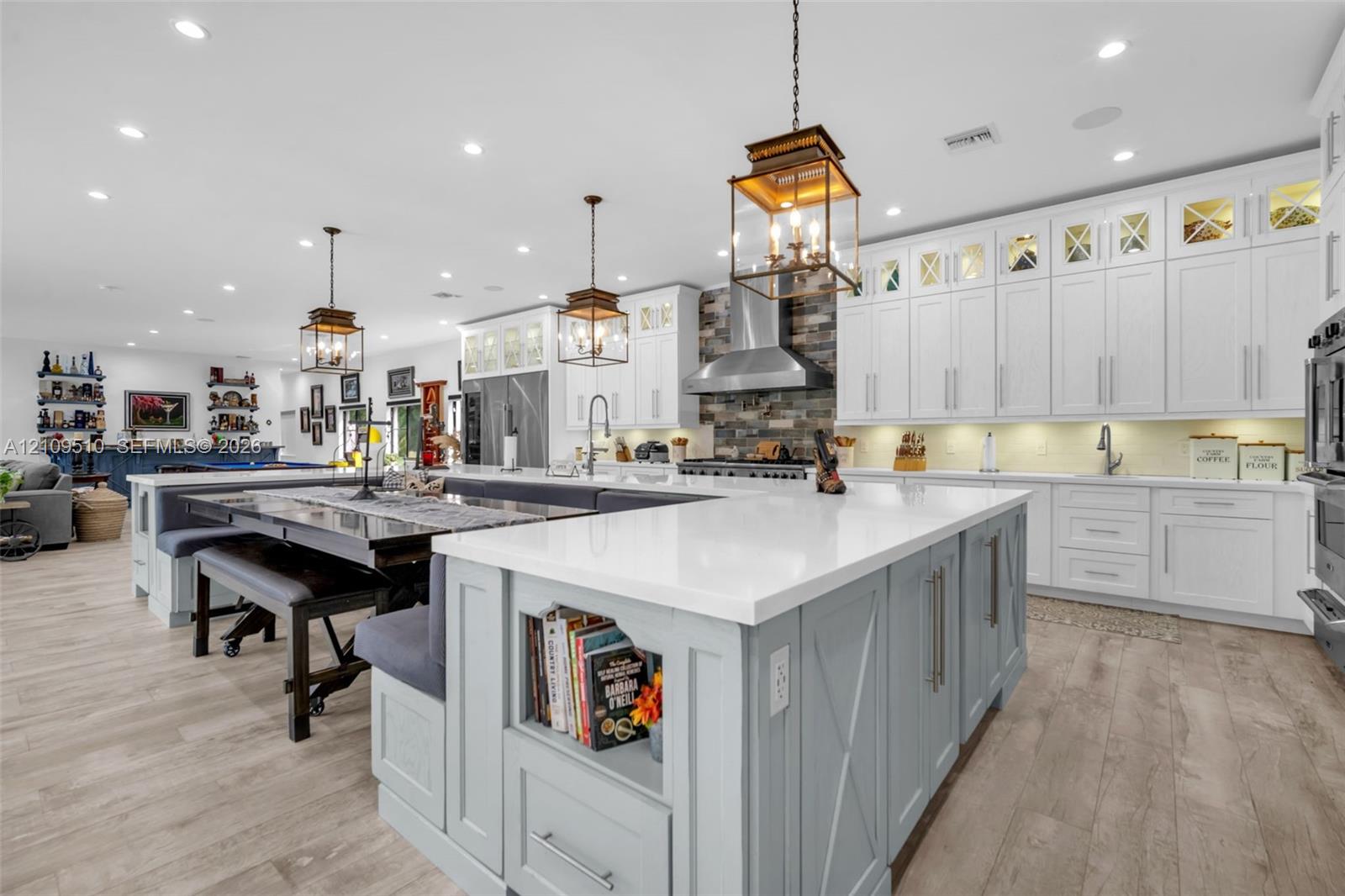 Kitchen featuring shaker-style cabinetry with glass-panel upper doors, a large central island with integrated storage, white quartz countertops, wood-finish flooring, and a stone-tile backsplash behind a stainless steel range hood
