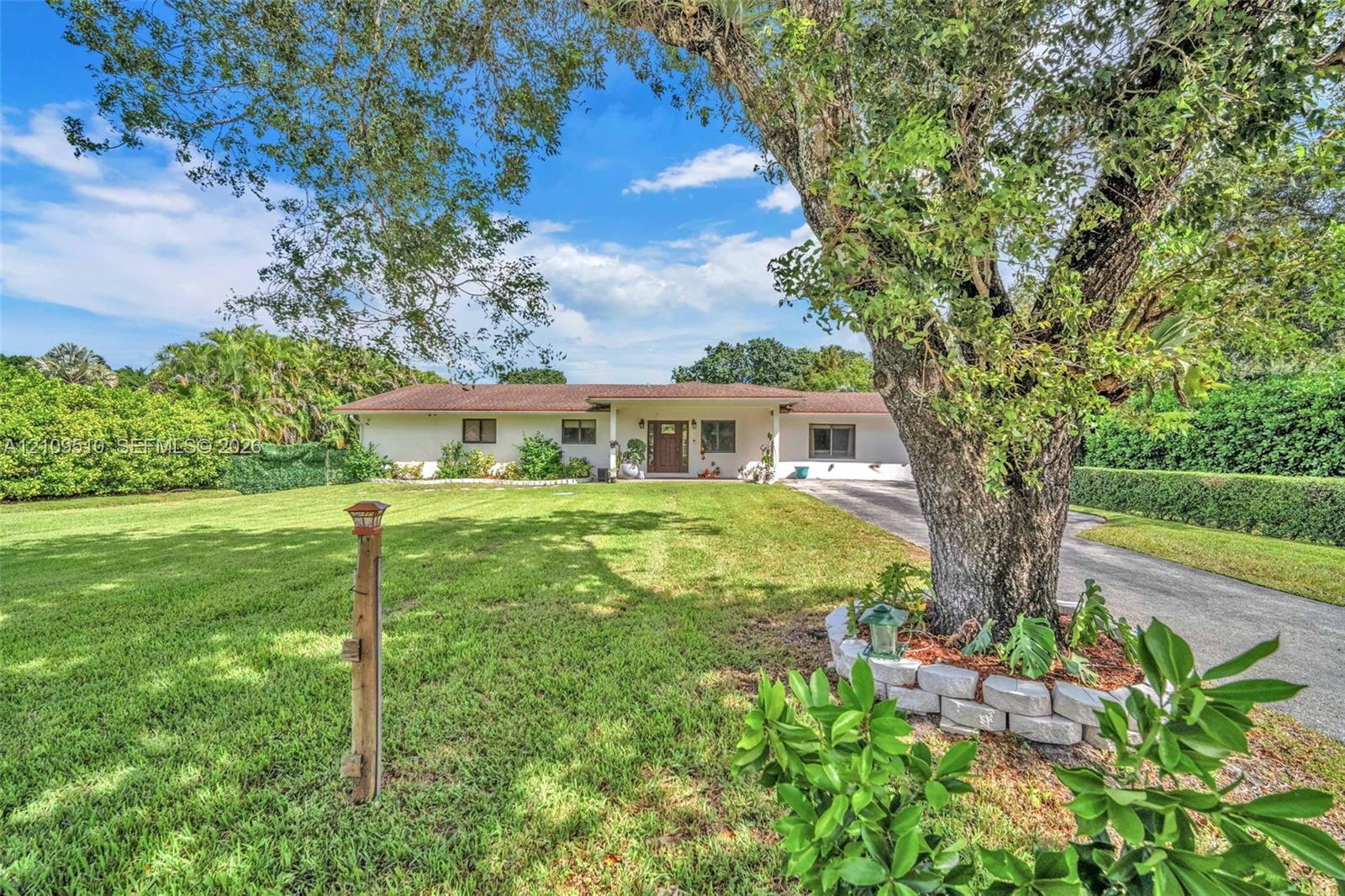 Single-story exterior featuring a light-toned stucco faade, a multi-paneled front entry door, expansive lawn, mature tree, and a concrete paver retaining wall