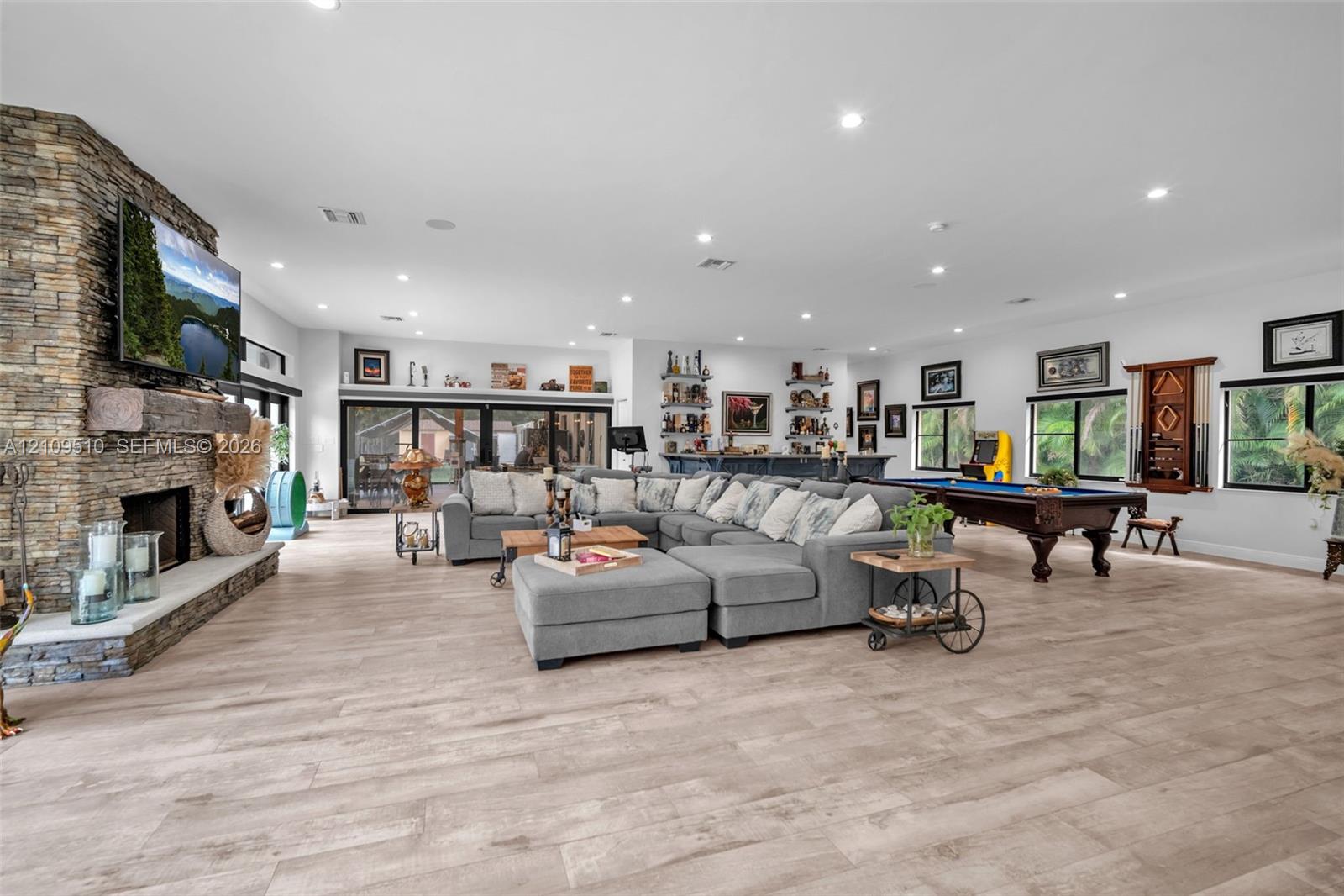 Open-concept living space featuring wood-finish flooring, floor-to-ceiling stone fireplace with a timber mantel, recessed lighting, and a built-in wet bar