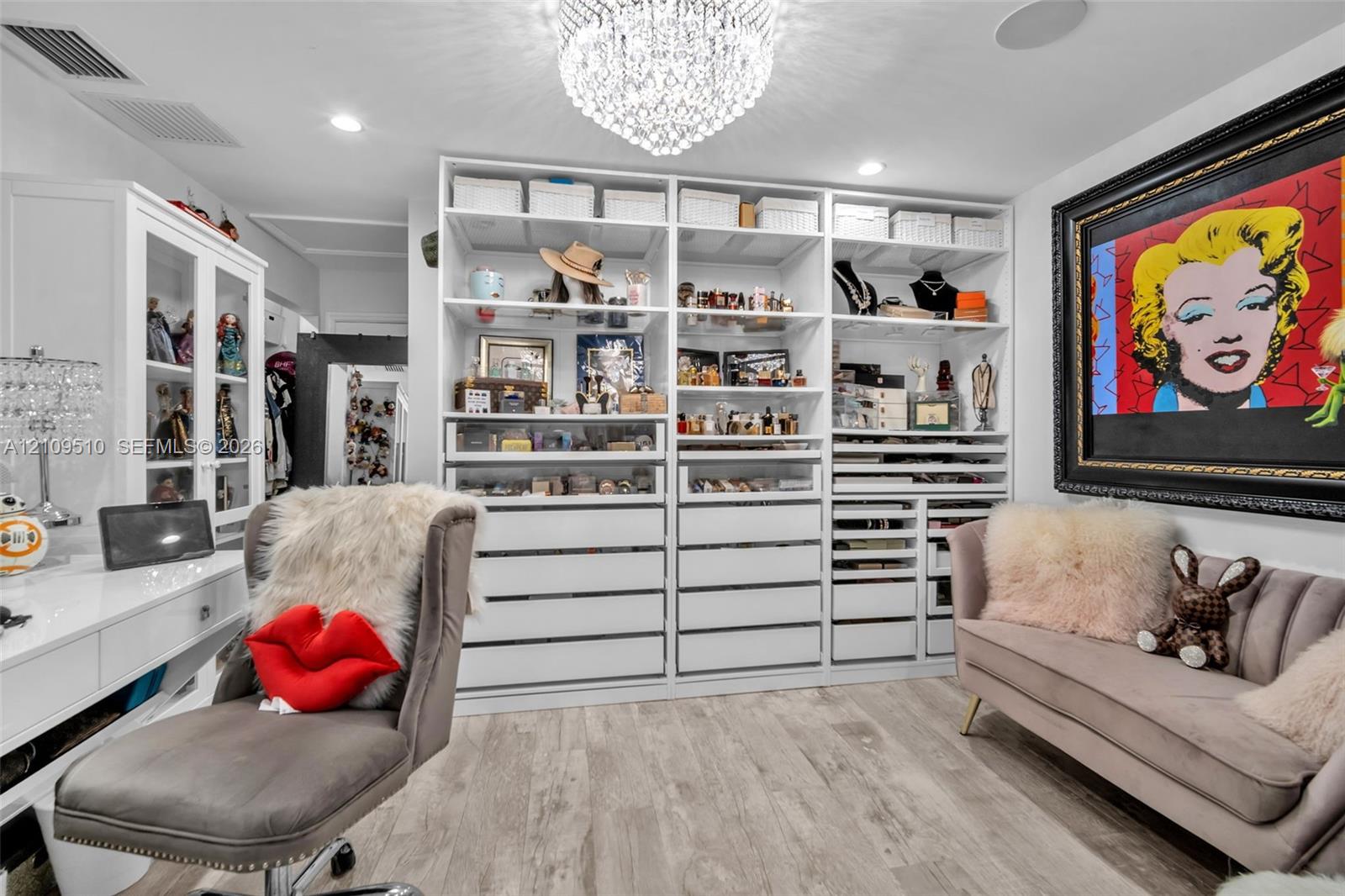 Custom built-in white cabinetry featuring extensive shelving, drawer storage, and glass-front display sections, complemented by wood-finish flooring and a crystal chandelier