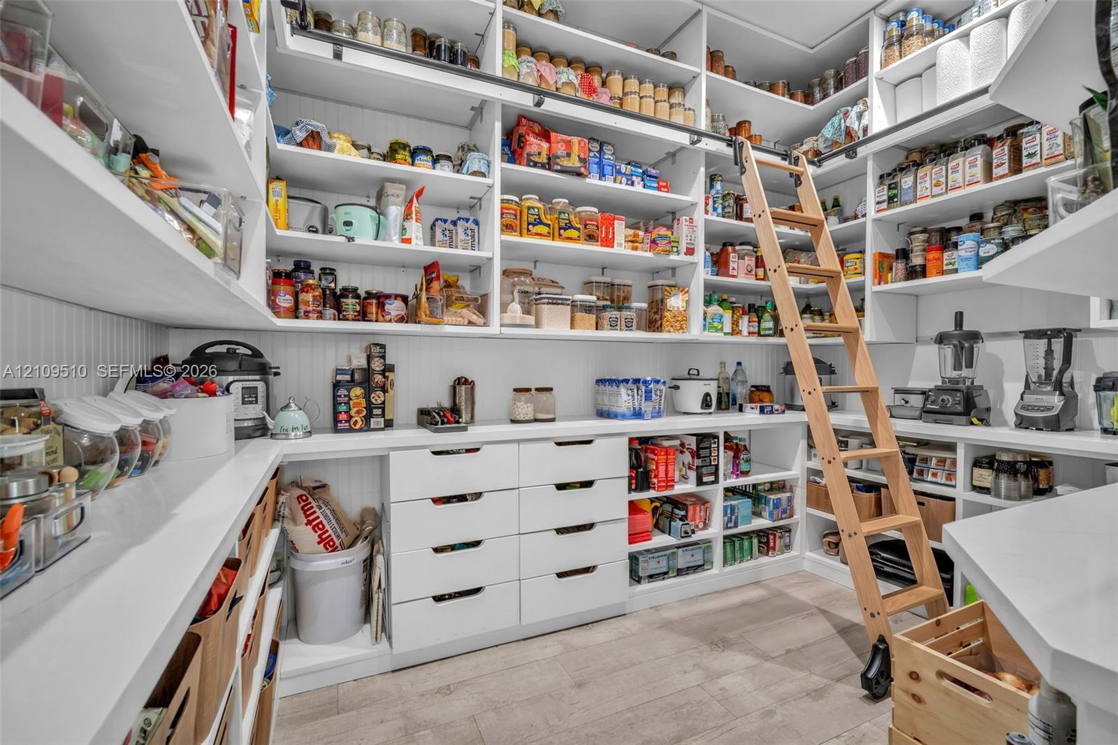 Custom walk-in pantry featuring expansive wall-to-wall shelving, floor-to-ceiling built-in cabinetry with integrated drawers, rolling library-style ladder, beadboard paneling, and wood-finish flooring
