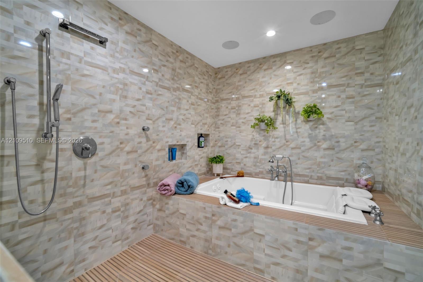 Walk-in wet room featuring textured tile walls, a deep soaking tub with chrome hardware, an integrated shower system with a handheld sprayer, built-in recessed wall shelving, and slatted wood-finish flooring