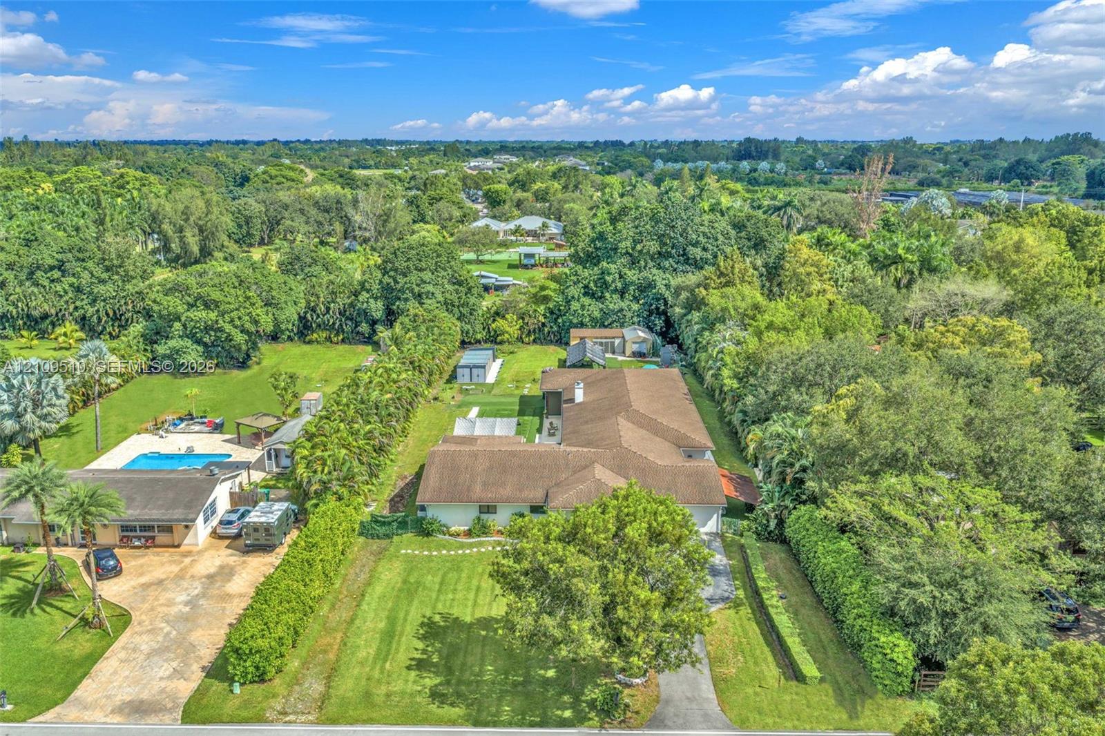 Aerial view of a sprawling property featuring an expansive lawn, mature tree canopy, multi-structured roofline, and an outdoor swimming pool with hardscaped surround