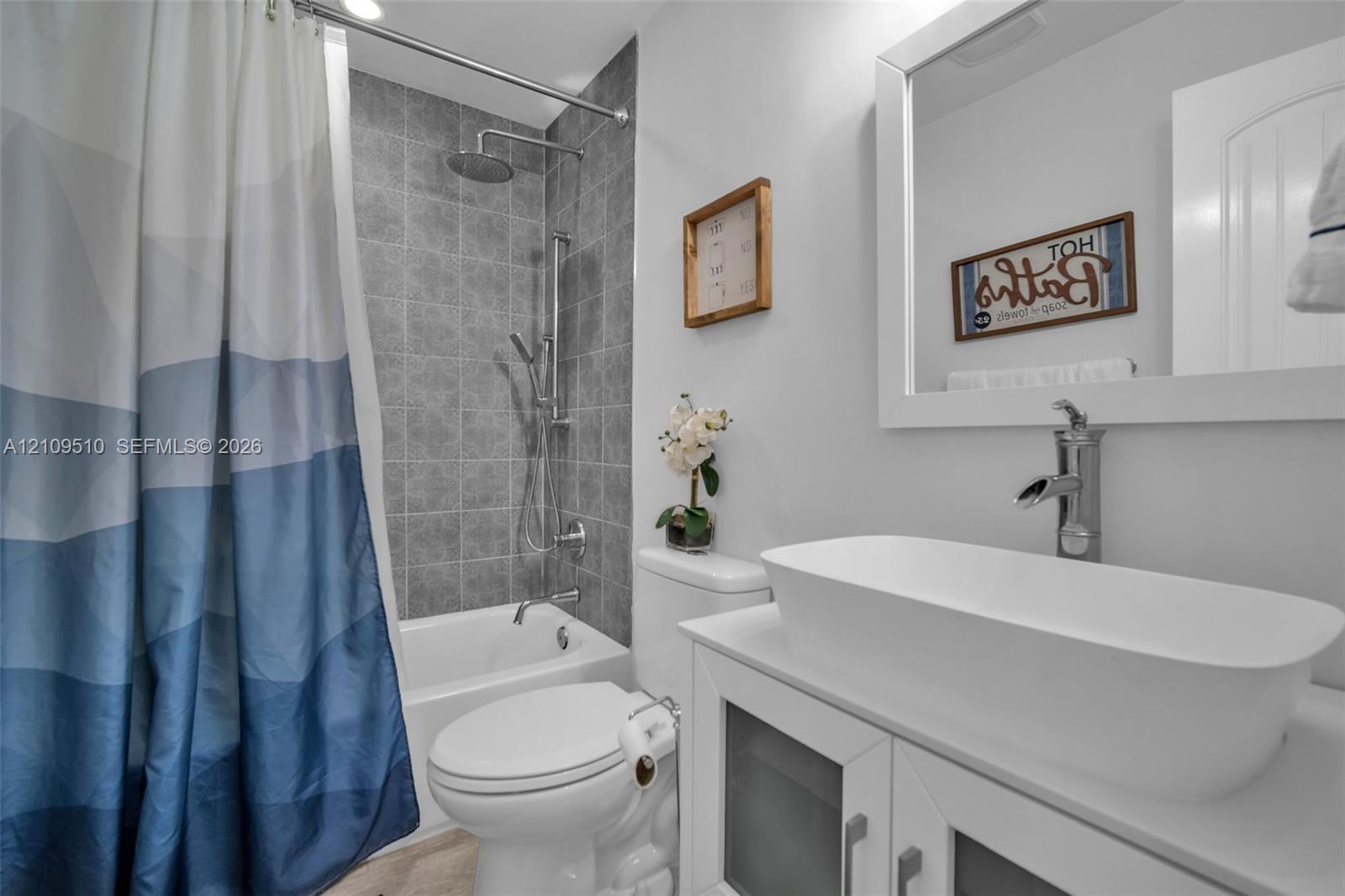 Bathroom featuring a white vessel sink with a chrome faucet, wall-mounted mirror with a white frame, shower with grey tile surround, adjustable handheld shower head, and vanity cabinet with frosted glass panel inserts