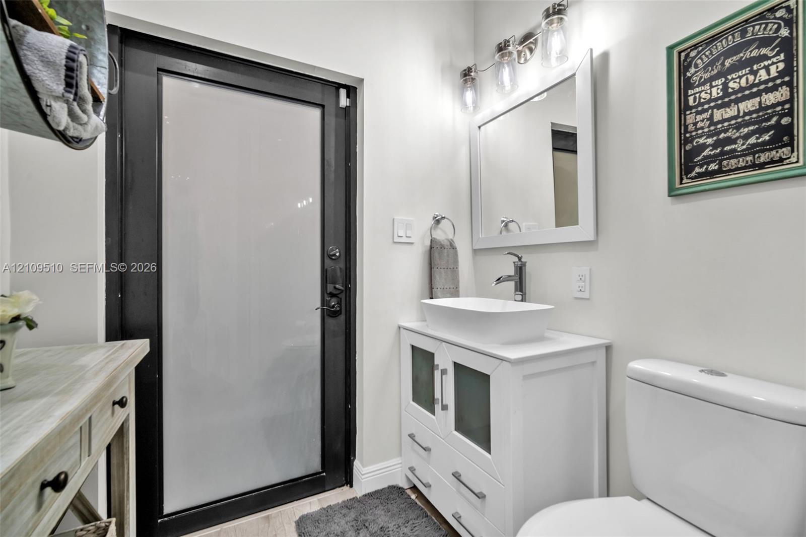 White vanity featuring a rectangular vessel sink, frosted glass cabinet inserts, and brushed nickel hardware, paired with a multi-light fixture, framed mirror, and a frosted glass door with a black frame