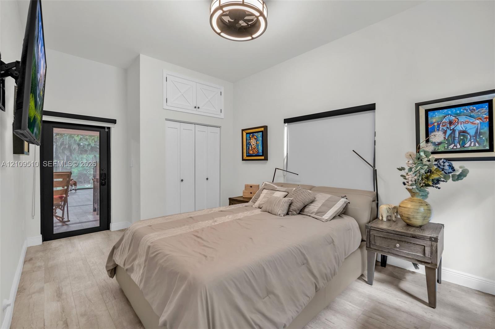 Neutral-toned room featuring wood-finish flooring, a contemporary drum-style ceiling light fixture, built-in double-door closet with overhead cabinet storage, and a black-framed glass door