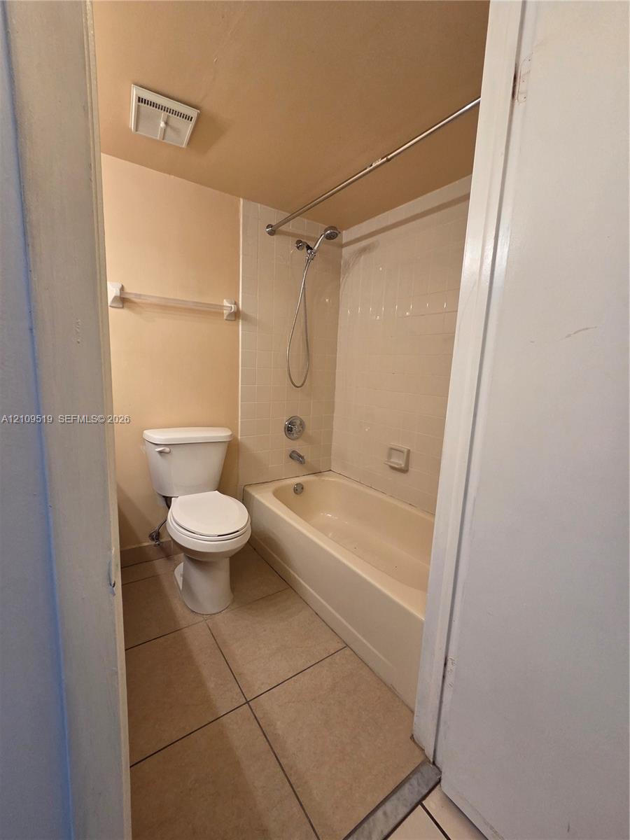 Bathroom featuring a porcelain toilet, a bathtub with a wall-mounted handheld shower fixture, tiled shower surround, and ceramic tile flooring