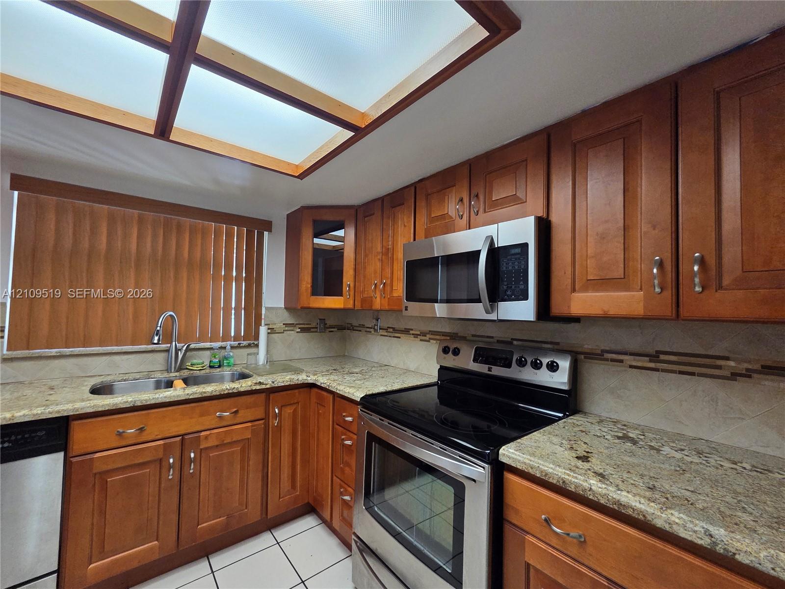 Kitchen featuring stained wood cabinetry, stone-surface countertops, built-in stainless steel appliances, a tile backsplash, and a recessed ceiling skylight