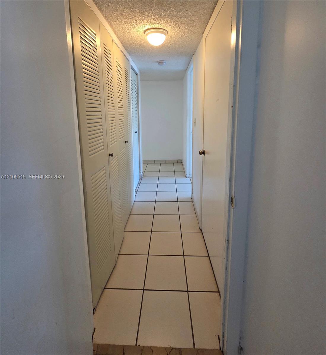 Hallway featuring light-colored tile flooring, multiple louvered-style closet doors, solid-panel doors, and a ceiling-mounted light fixture