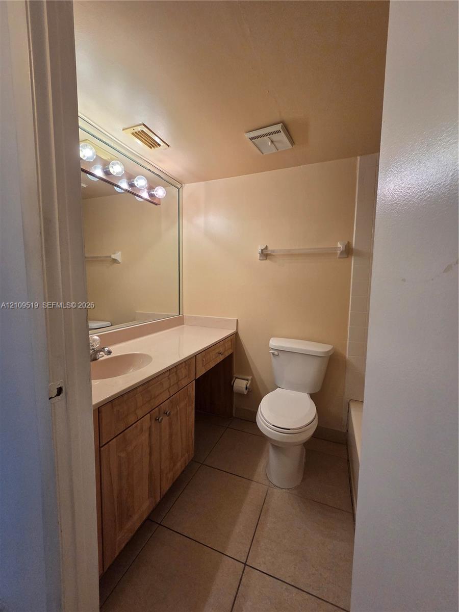 Bathroom featuring a vanity with wood-finish cabinetry, a solid surface integrated countertop, a full-width mirror with multi-bulb light fixture, and tile flooring