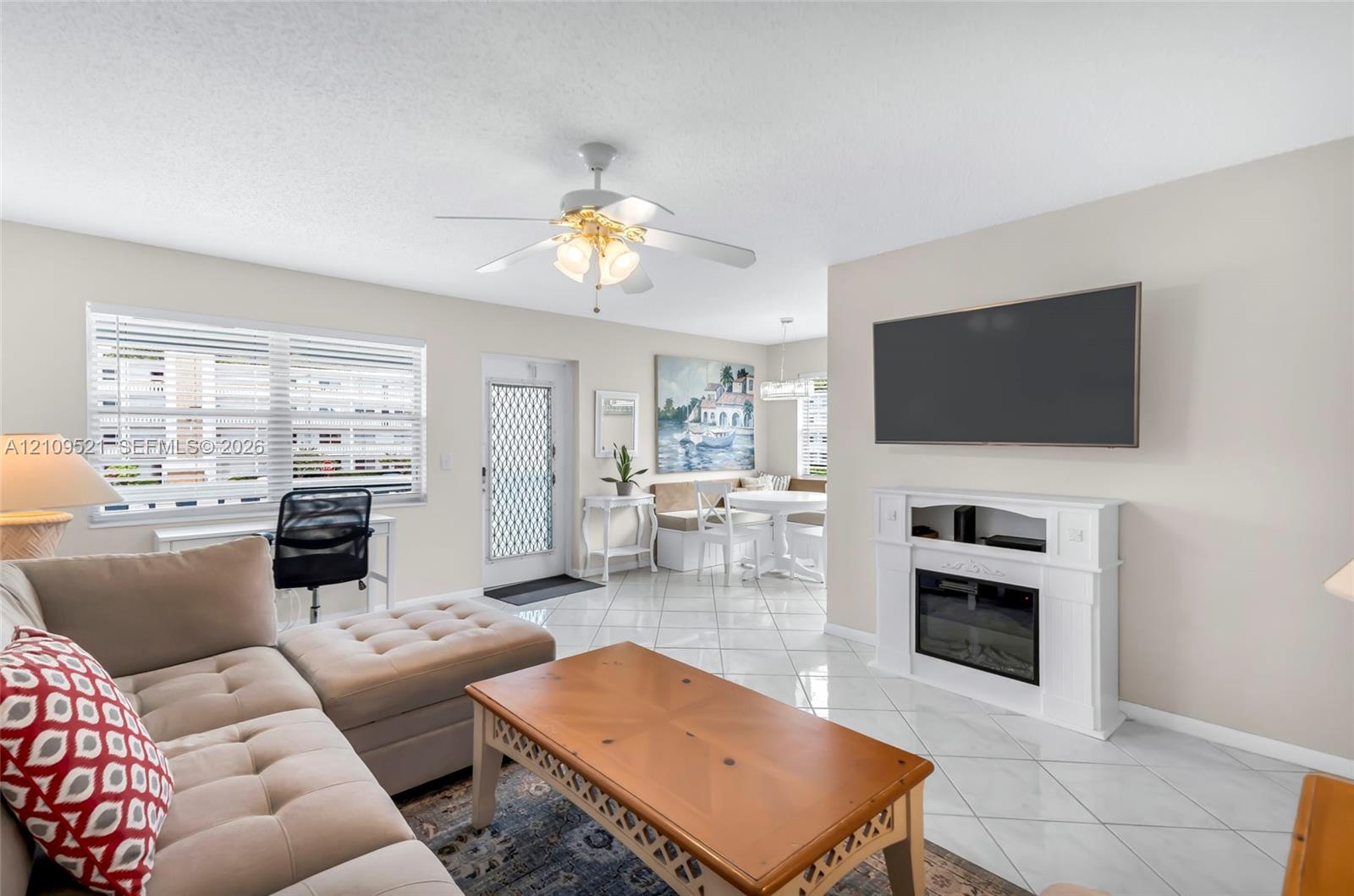 Open-concept living space featuring high-gloss tile flooring, white built-in electric fireplace, ceiling fan with integrated lighting, and white-framed exterior door with screen