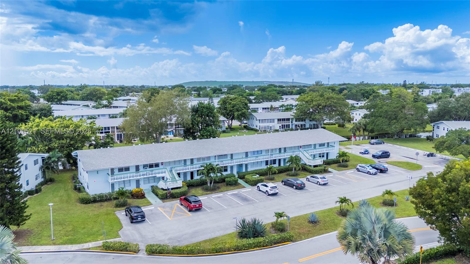 Multi-story residential building featuring a shingled roof, exterior balconies, paved surface parking, and manicured landscaping