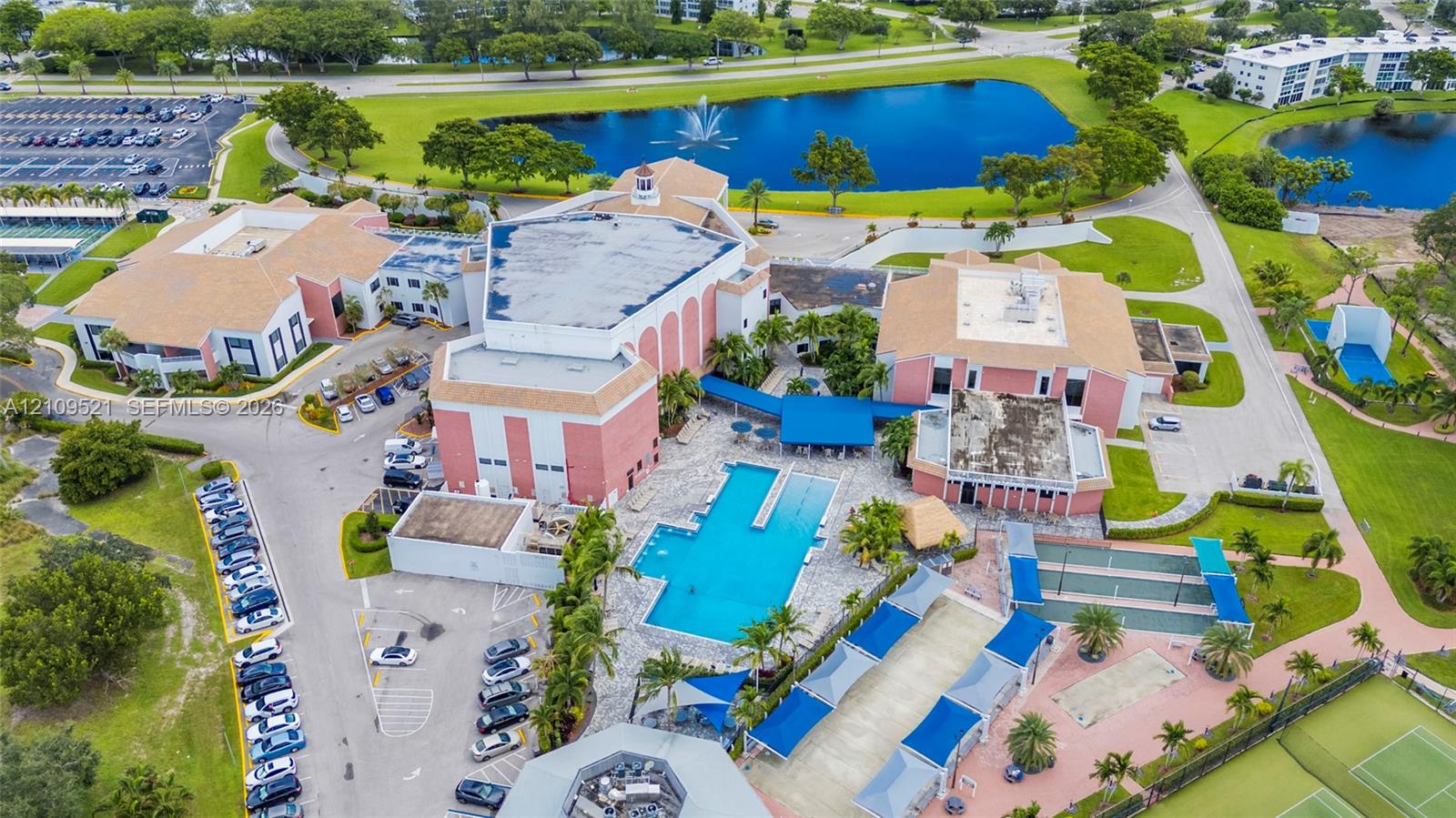 Aerial perspective featuring a multi-building property complex with a large swimming pool, paved surface parking areas, multi-court recreational facilities, and manicured grounds centered around a water feature with a fountain