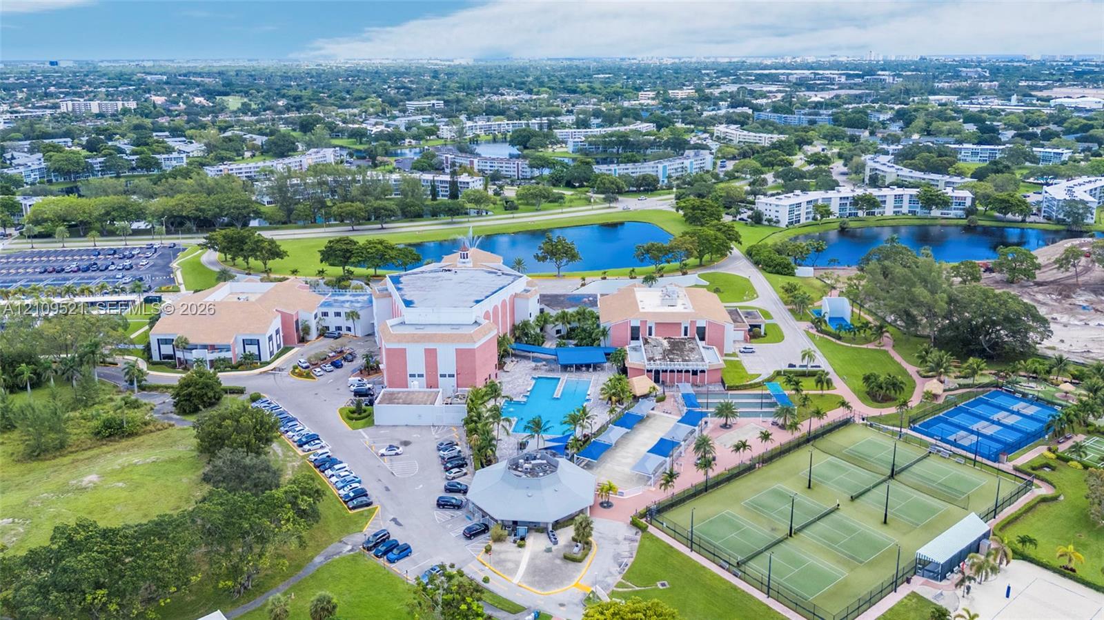 Aerial perspective of the complex featuring a multi-building facility, an outdoor swimming pool, multiple tennis courts, and landscaped green space surrounding a lake