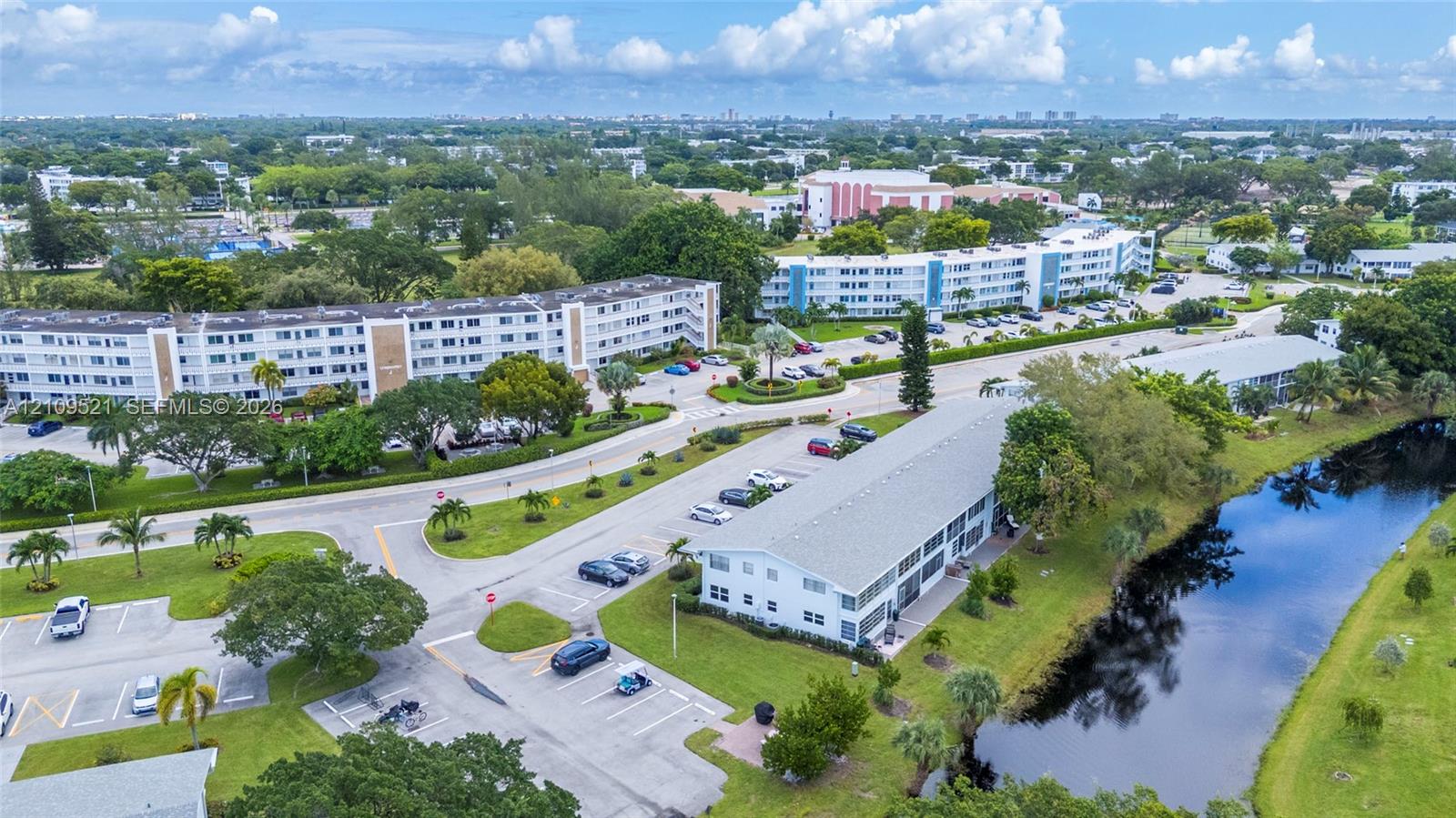 Aerial perspective featuring multi-story residential buildings, paved surface parking areas, manicured green spaces, and a bordering waterway