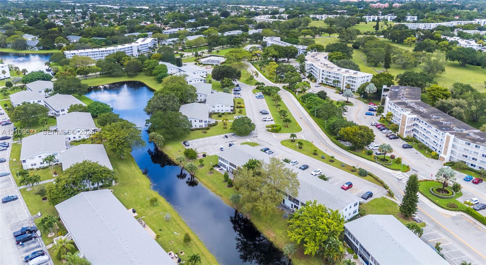Aerial perspective of residential complex featuring multi-story buildings, paved surface parking, mature landscaping, and an elongated water feature