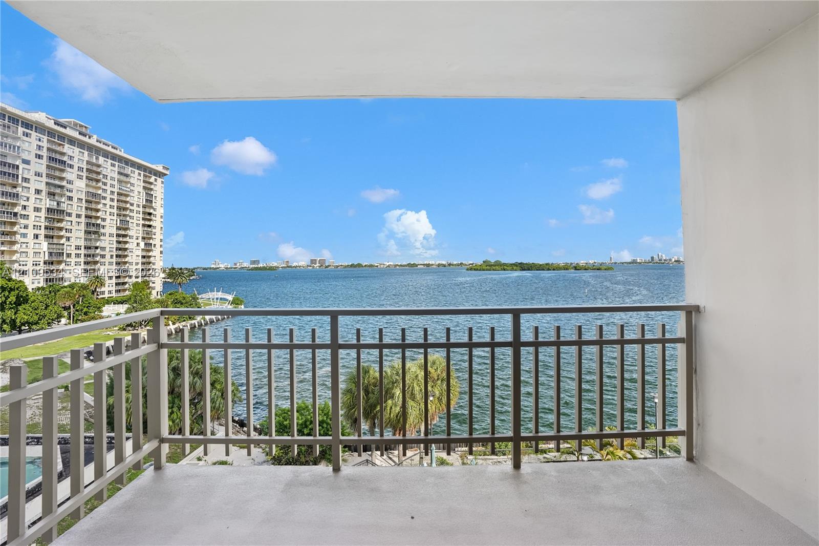 Covered balcony featuring metal railings and concrete flooring, overlooking expansive water scenery with coastal surroundings