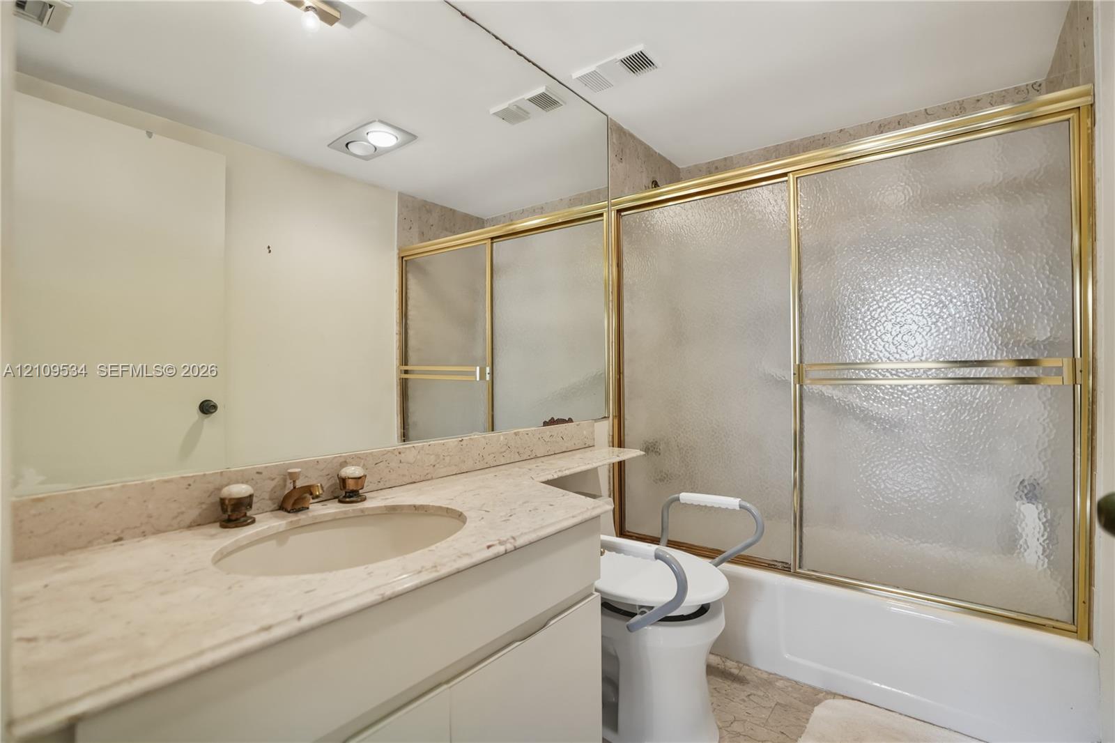 Stone-surface vanity countertop with an integrated oval sink, full-width wall mirror, and tub-shower combination featuring textured glass sliding doors with gold-tone frames