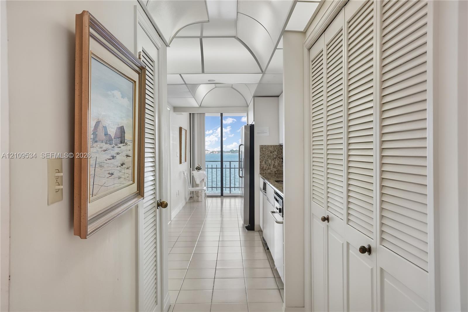 Hallway featuring white tile flooring, louvered closet doors, and a vaulted ceiling, leading to a balcony with water scenery