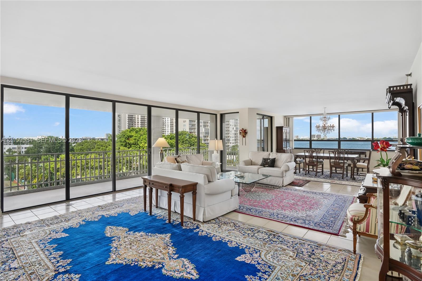 Expansive living area featuring floor-to-ceiling windows, tile flooring, a balcony with metal railing, and decorative chandelier lighting Expansive living area featuring floor-to-ceiling windows, tile flooring, a balcony with metal railing, and decorative chandelier lighting