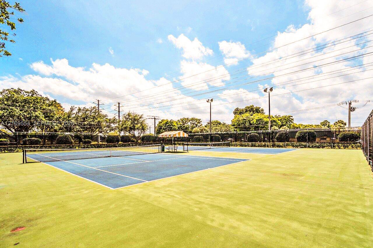 Multi-court tennis facility featuring blue hard-court surfaces, perimeter fencing, overhead lighting fixtures, and a shaded central seating area