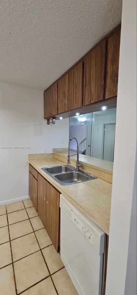 Kitchen featuring wood-finish cabinetry, neutral solid-surface countertops, dual-basin stainless steel sink with high-arch faucet, integrated dishwasher, and tiled flooring