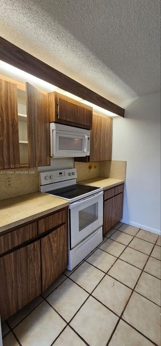 Kitchen featuring wood-finish cabinetry, white electric range with matching over-the-range microwave, solid-surface countertops, and ceramic tile flooring