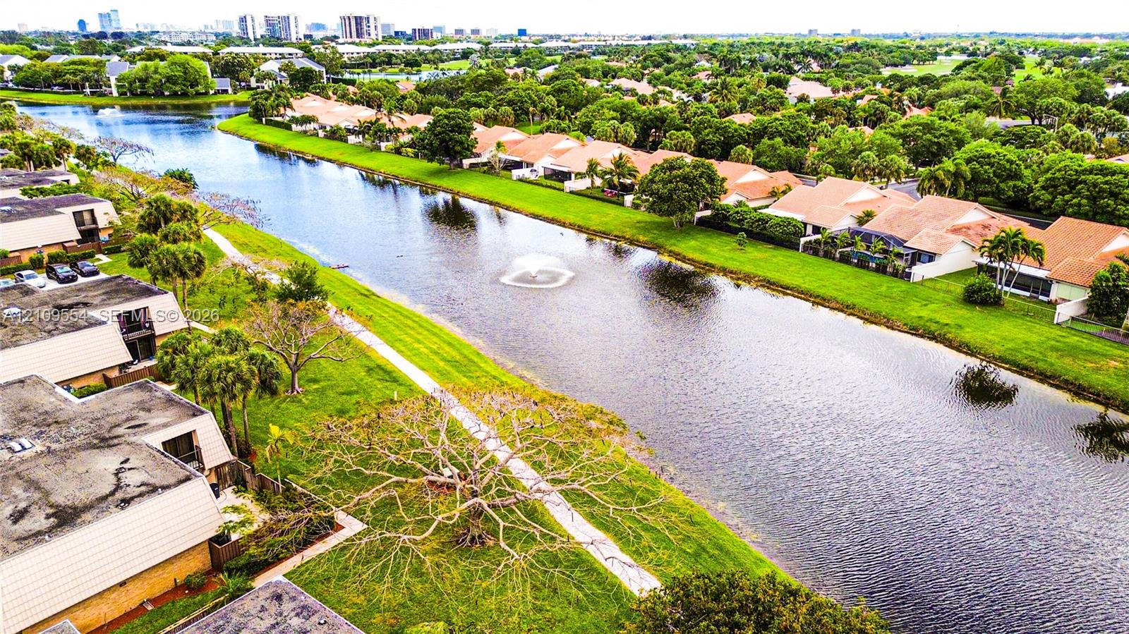 Aerial perspective of a multi-unit residential community featuring a central canal, water fountain, paved walkways, and mature landscaping