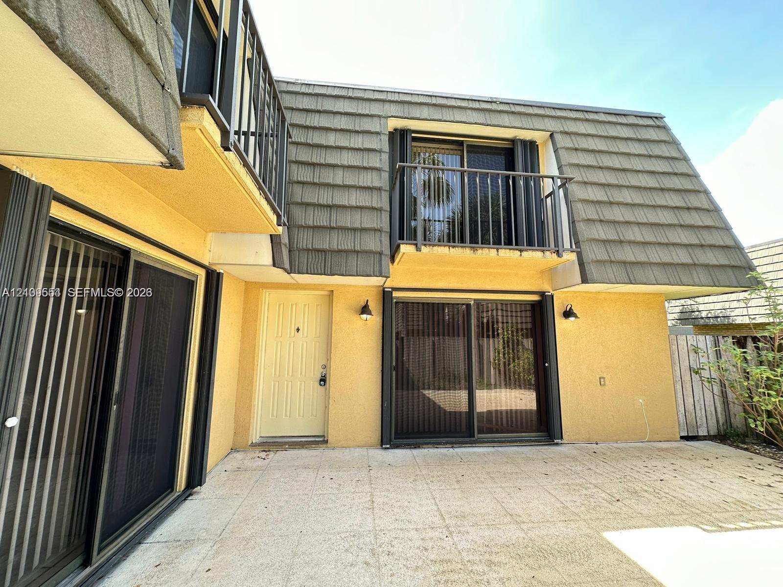 Exterior facade featuring a second-story balcony with metal railings, sliding glass patio doors, shingled mansard roof design, and a concrete patio