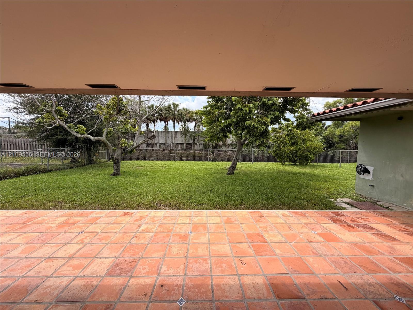 Covered patio featuring terracotta tile flooring, overlooking a backyard with mature trees, green lawn, and perimeter fencing