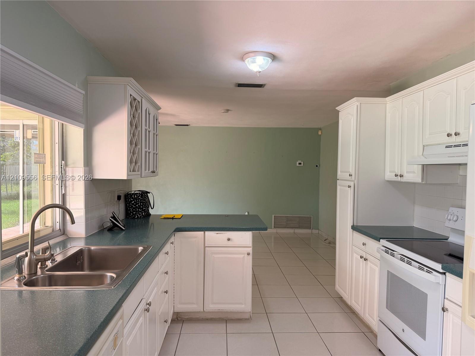 Kitchen featuring white cabinetry with glass-front detailing, solid surface countertops, dual-basin stainless steel sink with high-arc faucet, white electric range, and tile flooring