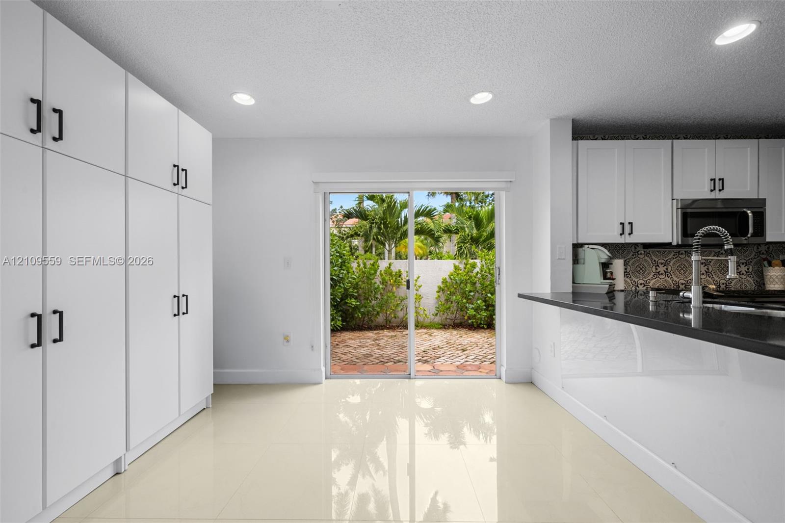 Kitchen featuring crisp white floor-to-ceiling cabinetry, dark stone countertops, patterned tile backsplash, polished neutral tile flooring, and sliding glass doors with exterior brick patio access