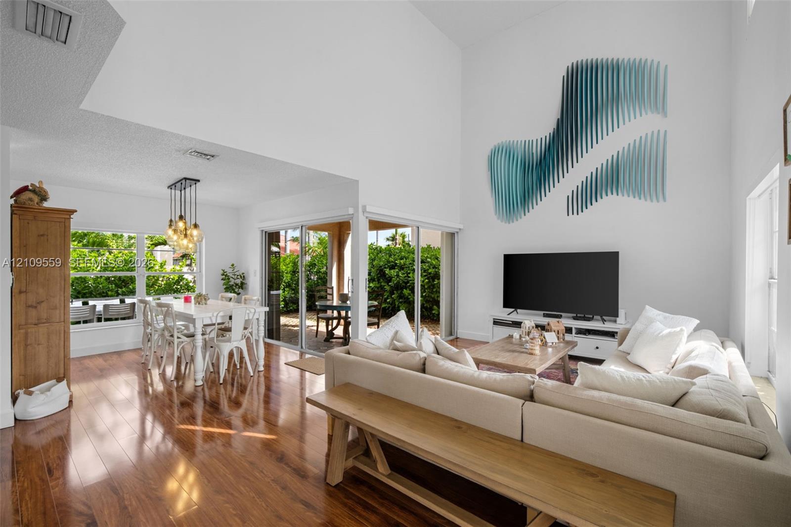 High-ceiling living area featuring wood-finish flooring, floor-to-ceiling glass sliding doors, and multi-pendant dining room lighting