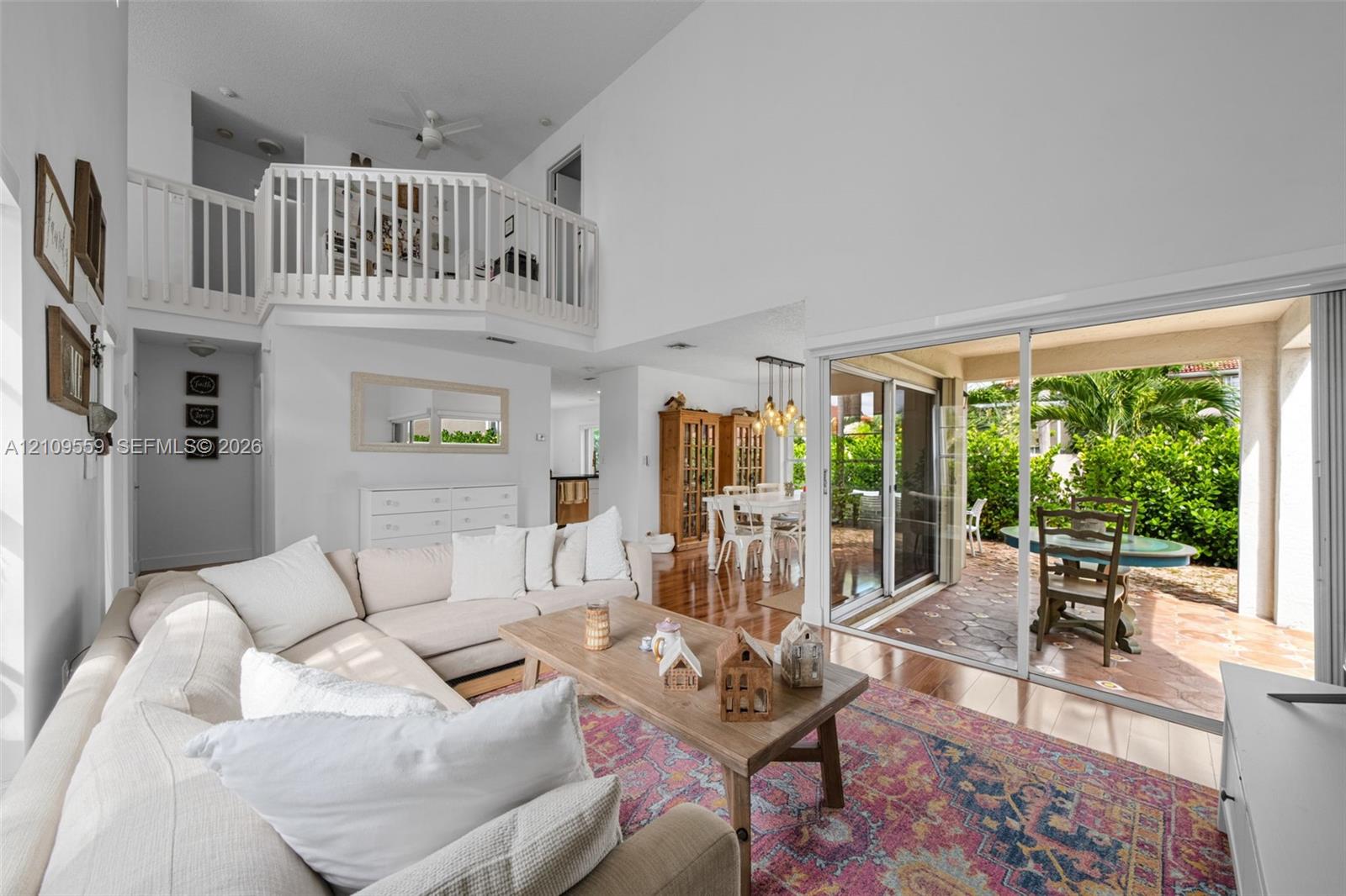 Living area with vaulted ceilings, elevated mezzanine loft with white railing, wood-finish flooring, and large sliding glass doors opening to a tiled patio