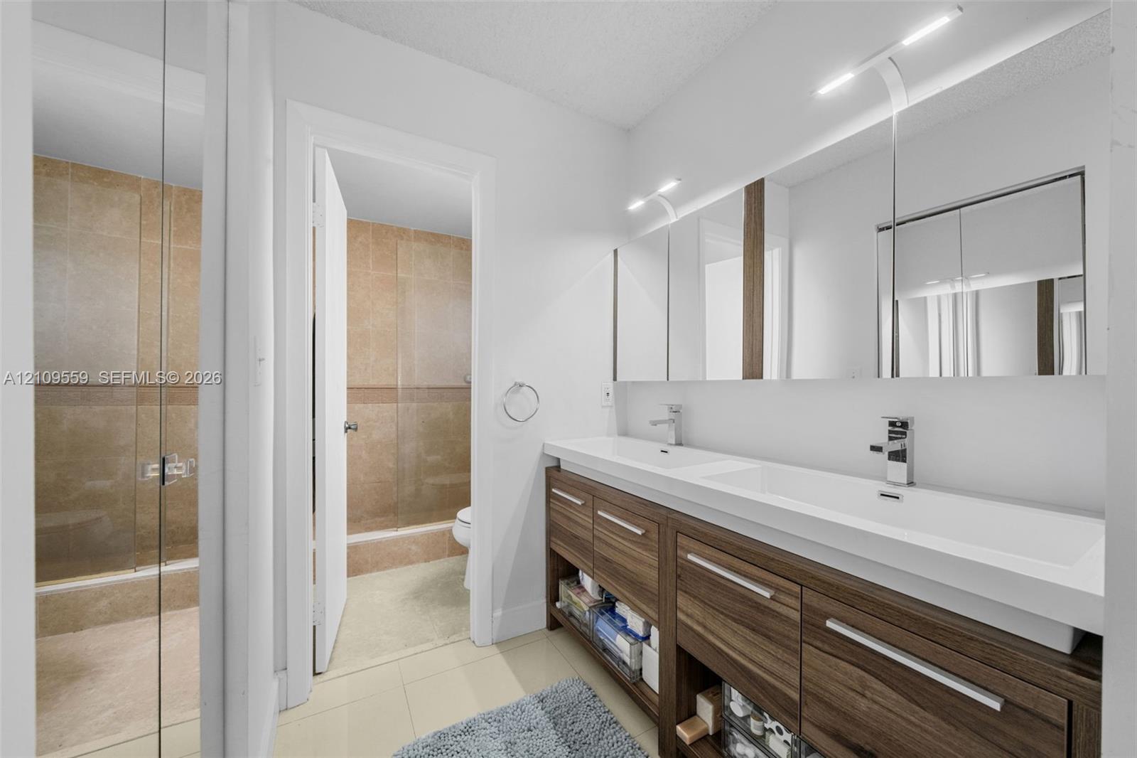 Dual-sink vanity featuring wood-finish cabinetry, integrated rectangular basins, and full-width mirror cabinet storage, complemented by a glass-enclosed shower with tile surround