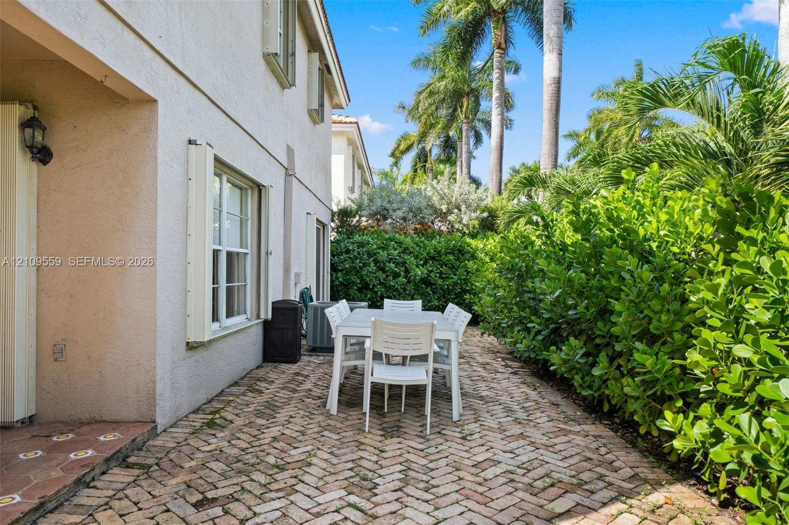 Herringbone-patterned brick paver patio featuring stucco exterior walls, multi-pane windows, and manicured perimeter hedges