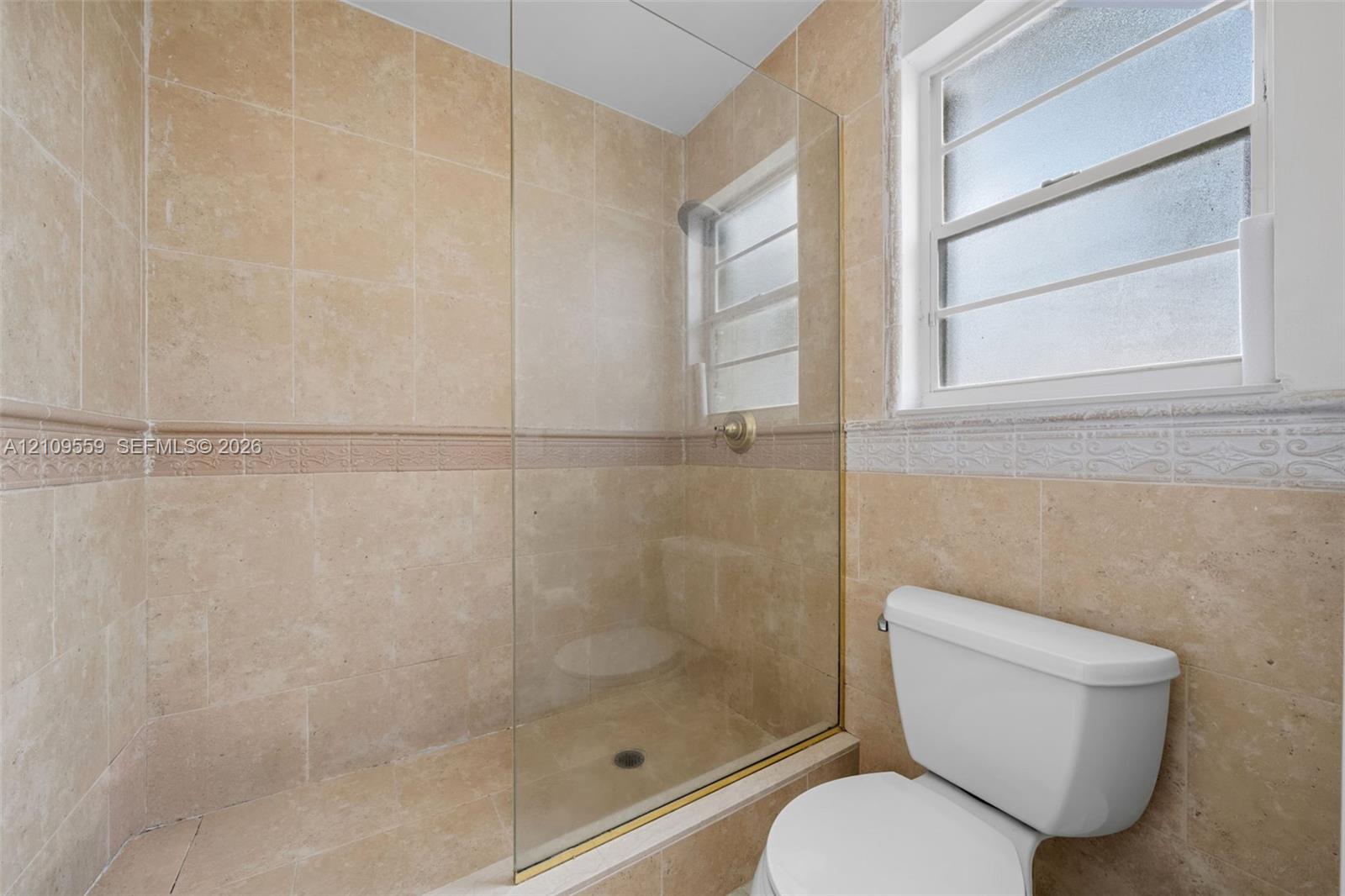 Full-height natural stone tiling with decorative mosaic accent border, frameless glass shower enclosure, brushed metal shower fixture, and louvered window