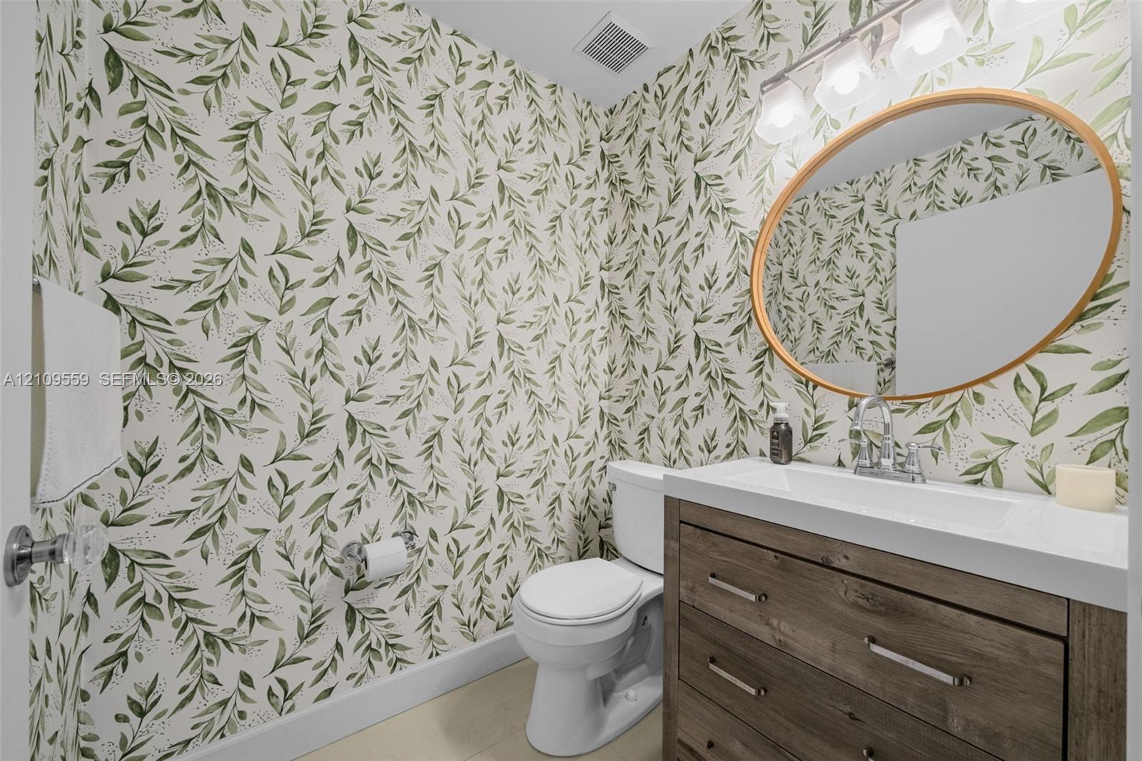Restroom featuring a wood-finish vanity, white rectangular countertop sink, oval gold-rimmed mirror, and botanical wallpaper