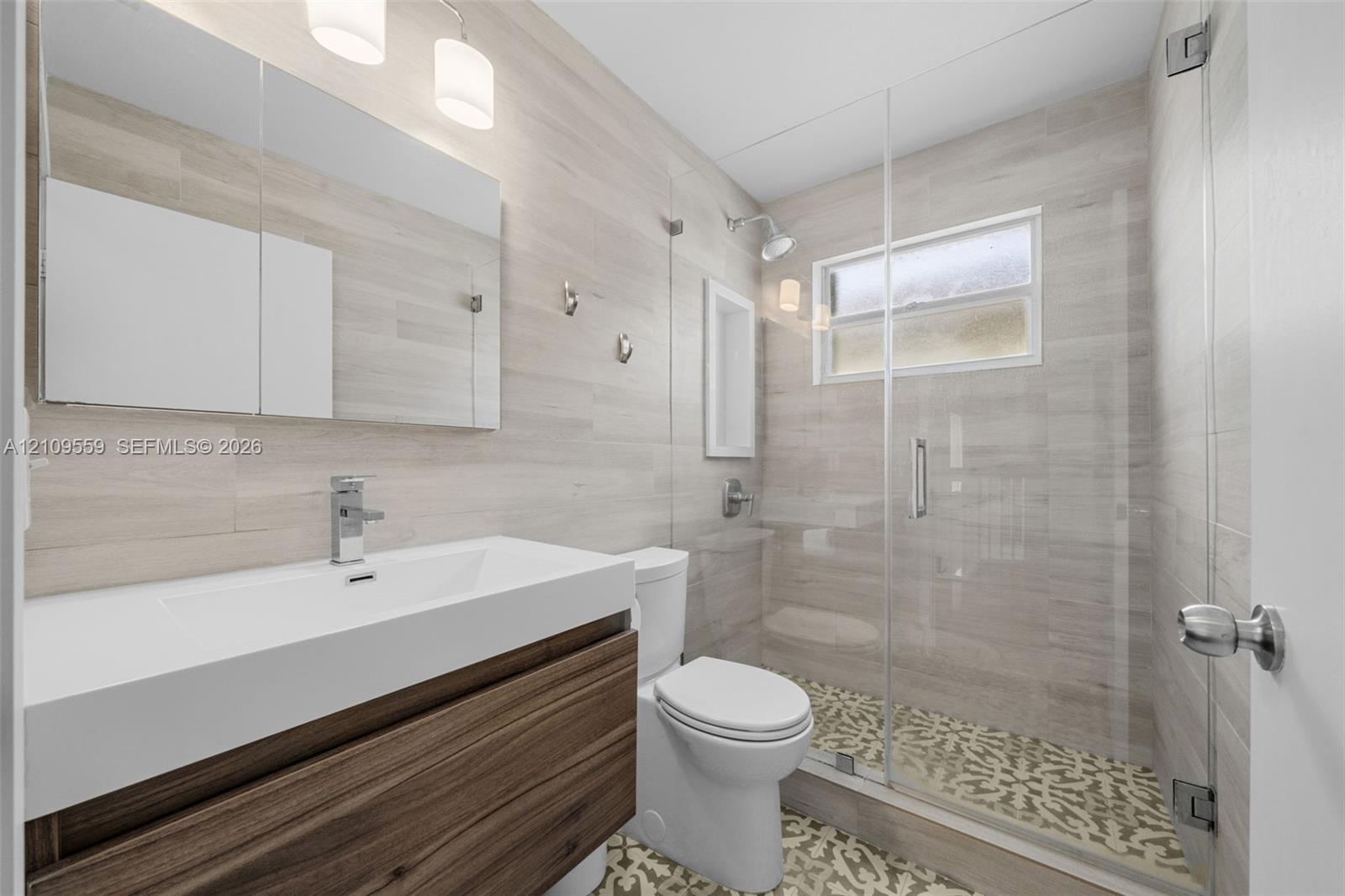 Floating vanity with a rectangular sink, wood-finish cabinetry, frameless glass shower enclosure, patterned tile shower flooring, and light-toned horizontal wall tile