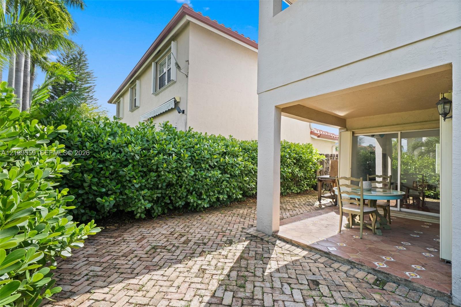 Covered patio with ceramic tile flooring, brick paver hardscaping, mature privacy hedges, and a wall-mounted exterior light fixture