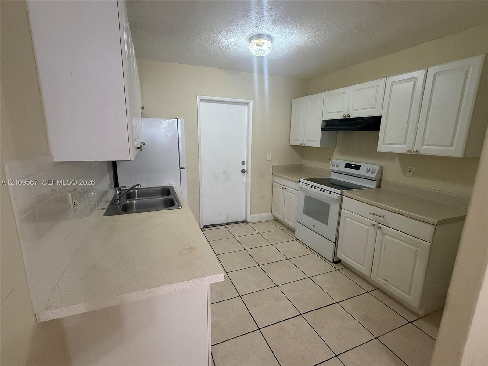 Kitchen featuring white cabinetry, solid surface countertops, stainless steel dual-basin sink, tile flooring, and integrated range hood Kitchen featuring white cabinetry, solid surface countertops, stainless steel dual-basin sink, tile flooring, and integrated range hood