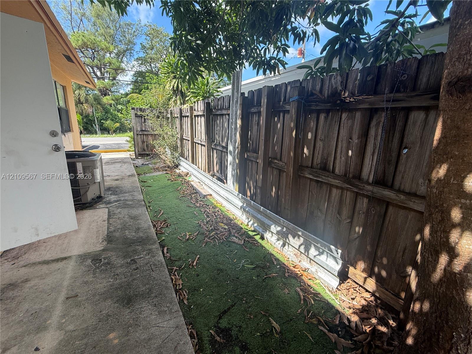Exterior side yard featuring concrete hardscaping, vertical wood privacy fencing with a metal base plate, and mature trees