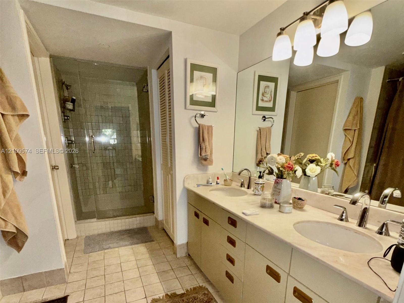 Double vanity featuring integrated sinks, light-toned countertop, and substantial cabinetry, complemented by a glass-enclosed shower with tiled surround and multi-light wall fixture Double vanity featuring integrated sinks, light-toned countertop, and substantial cabinetry, complemented by a glass-enclosed shower with tiled surround and multi-light wall fixture