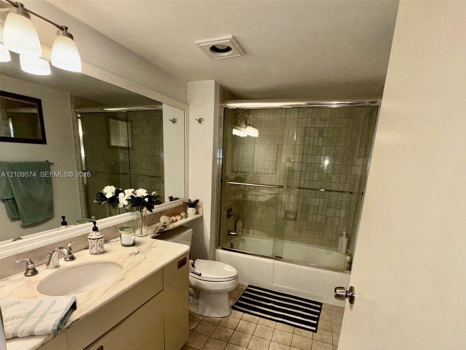 Full-width wall mirror, vanity cabinet with integrated sink, stone-finish countertop, shower and bathtub combination with sliding glass doors, and tiled flooring Full-width wall mirror, vanity cabinet with integrated sink, stone-finish countertop, shower and bathtub combination with sliding glass doors, and tiled flooring