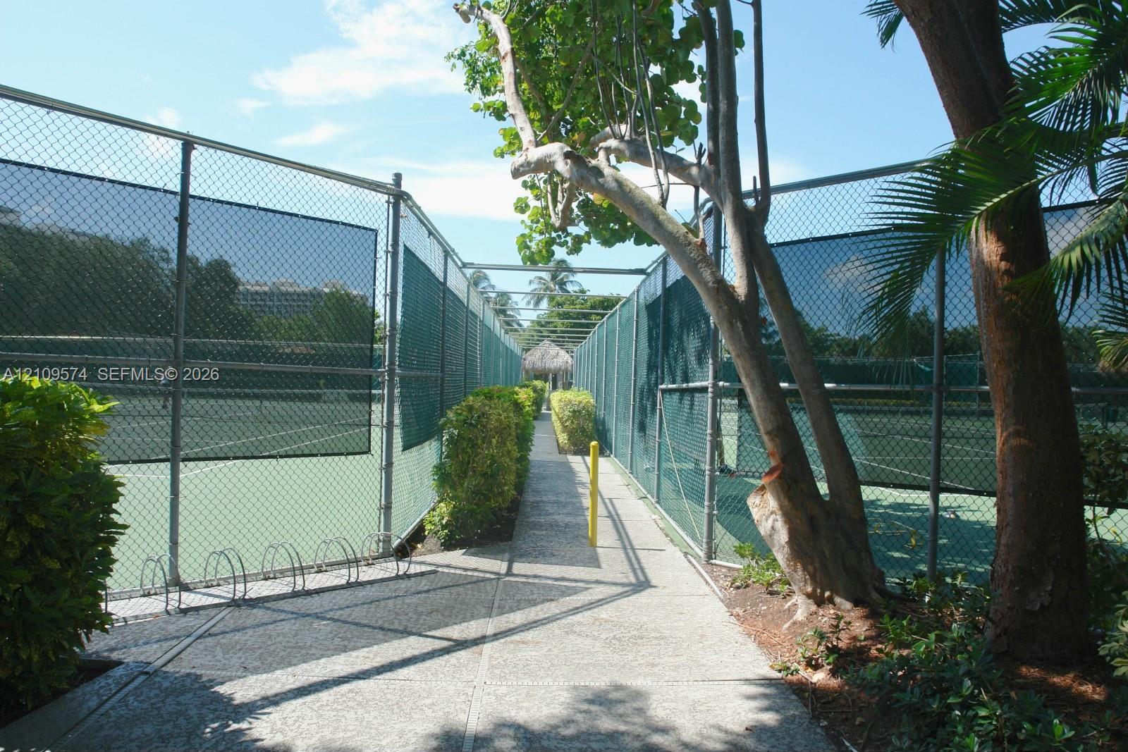 Tennis courts enclosed by high chain-link fencing, featuring paved walkways and mature tropical landscaping